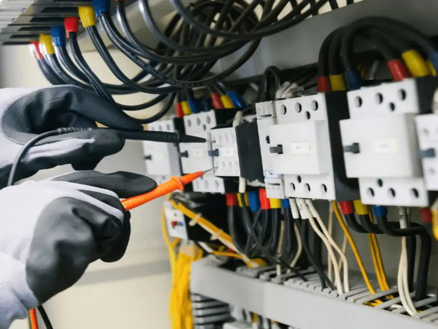 Electrician in insulated gloves testing wiring inside an electrical panel with a meter probe